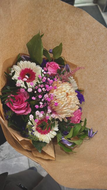 A vibrant floral bouquet featuring a mix of pink roses, white daisies with pink centers, cream-colored carnations, and small purple flowers, all arranged with green foliage. The flowers are wrapped in