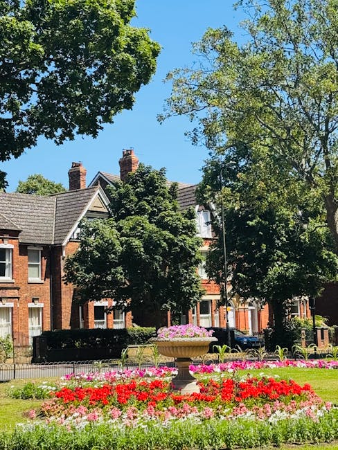 A vibrant garden scene with a well-maintained flower bed featuring red, pink, and white flowers arranged in a circular pattern around a central stone planter filled with pink flowers. Tall, leafy gree