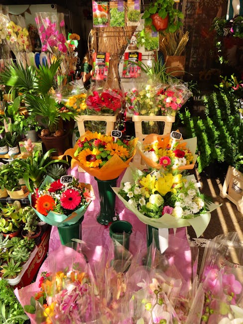 A vibrant display of fresh flower bouquets arranged on a pink cloth-covered table inside a florist shop. The bouquets feature a variety of colorful flowers, including bright pink gerbera daisies, yell
