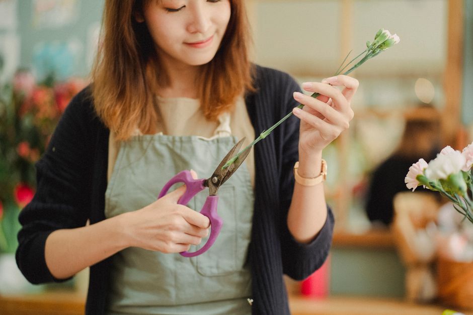 A young woman with shoulder-length brown hair, dressed in a beige top and a black cardigan, is in a floral shop, holding a pair of pink handled scissors in her right hand and a green stem with small w